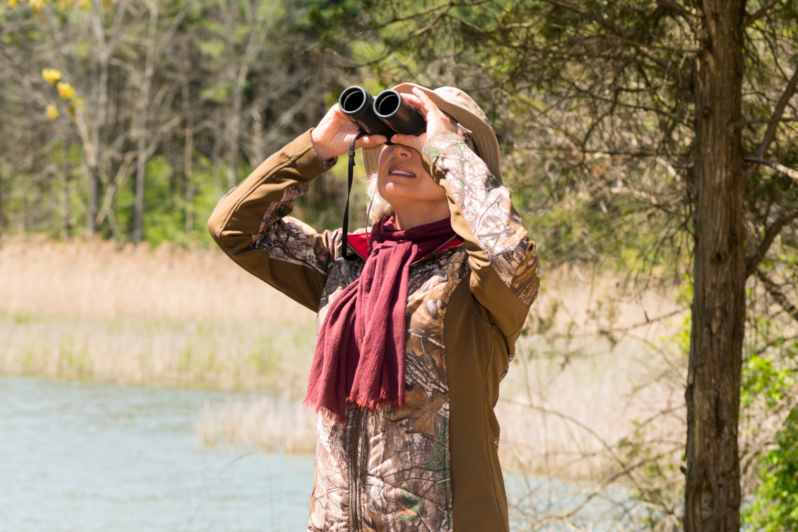 woman in outdoor clothing looking through binoculars outside near a pond